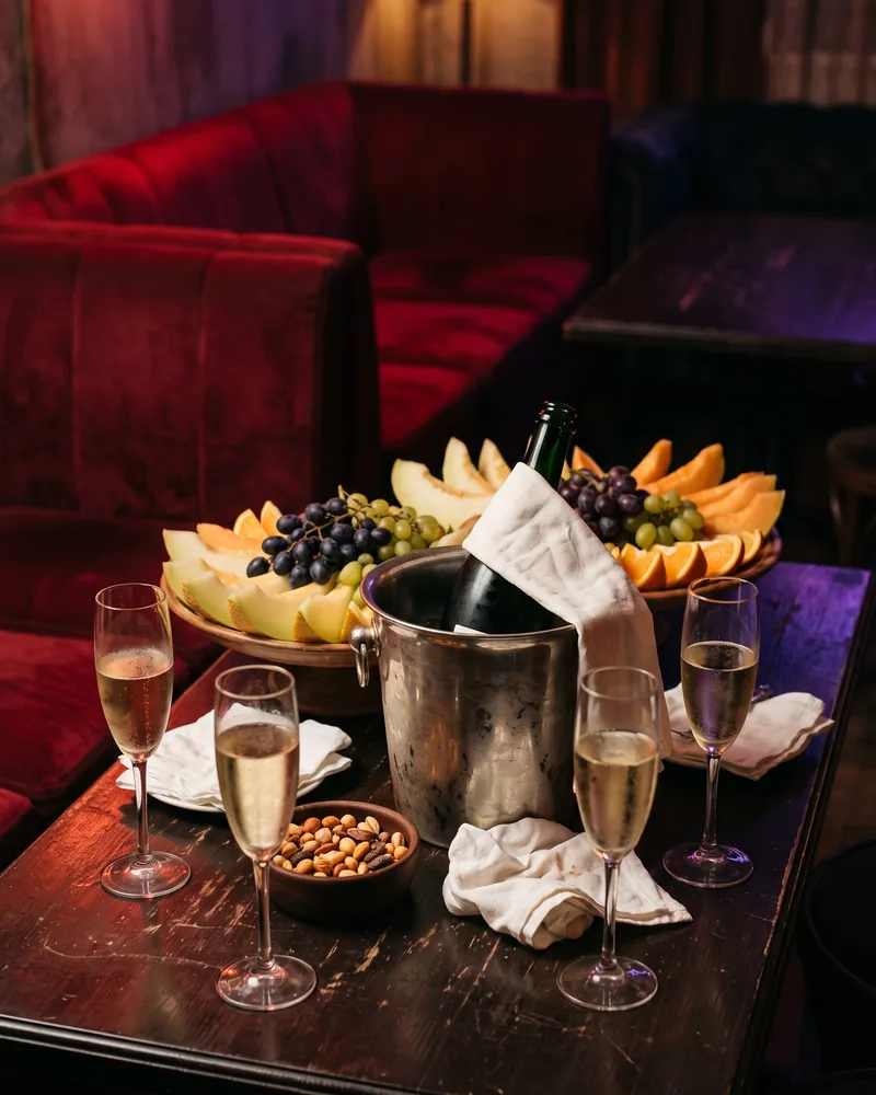 A lounge table beside red velvet banquettes with a bottle in an ice bucket, 4 flutes of sparkling wine, a bowl of nuts and a platter of sliced fruit