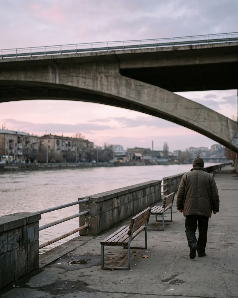 A concrete river embankment under a road bridge at dawn, with the river flat and grey, empty benches and one man walking away