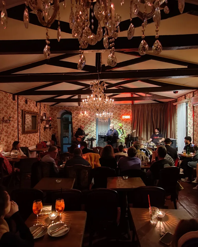 A band with guitars, a keyboard and drums playing at the end of a room with floral wallpaper, crystal chandeliers and a seated audience