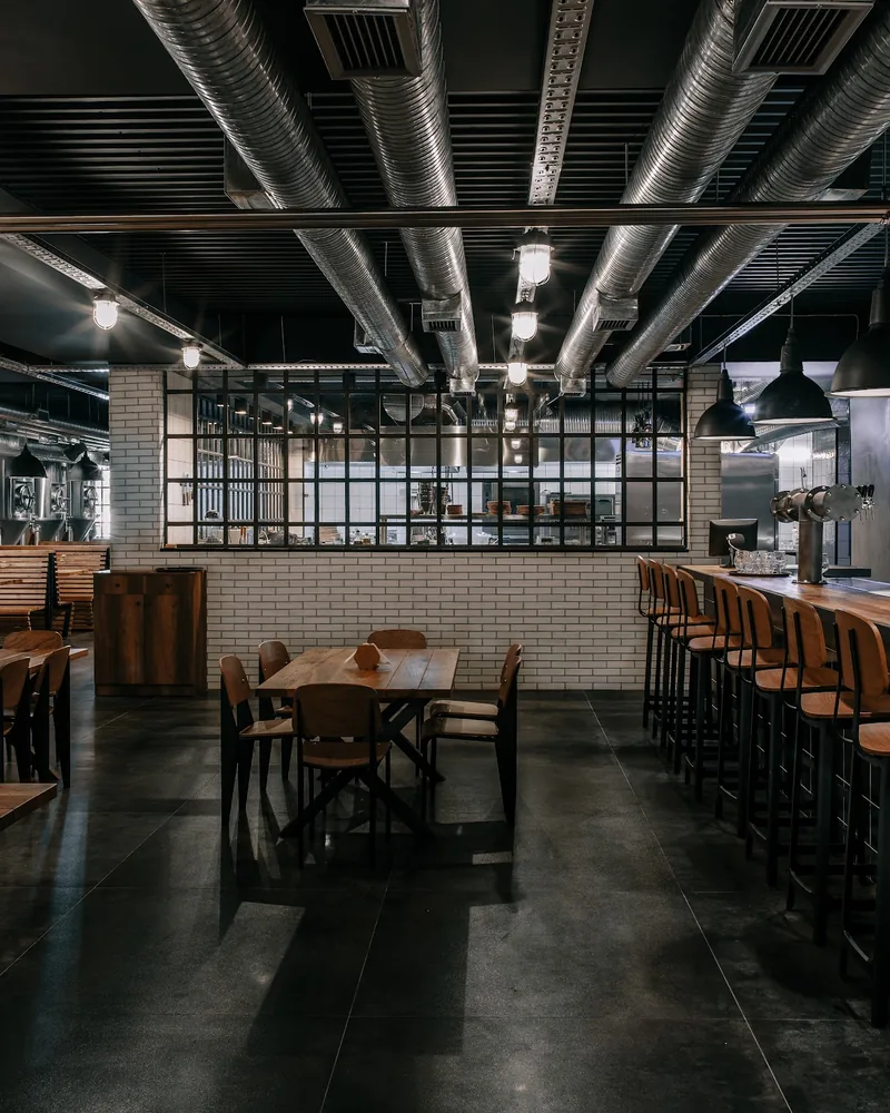 A taproom with wooden tables, a long bar with a tap tower on the right and steel brewing tanks behind glass at the back left