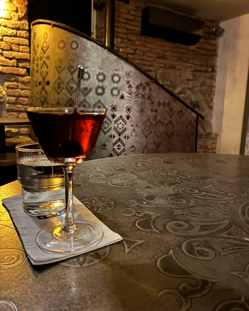 A dark red cocktail in a coupe with a cherry, beside a glass of water on a carved table at 41° Art of Drinks