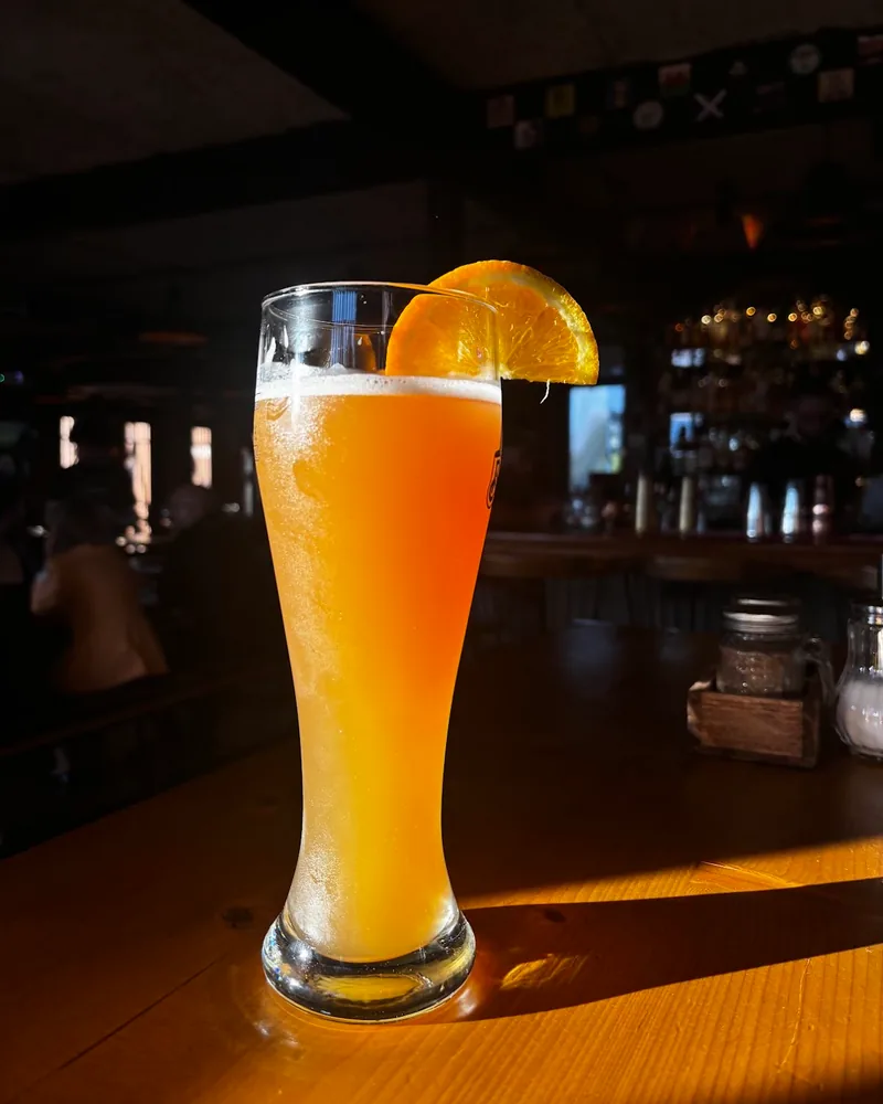 A tall glass of cloudy golden wheat beer with an orange slice on the rim, lit by low sun on a wooden bar