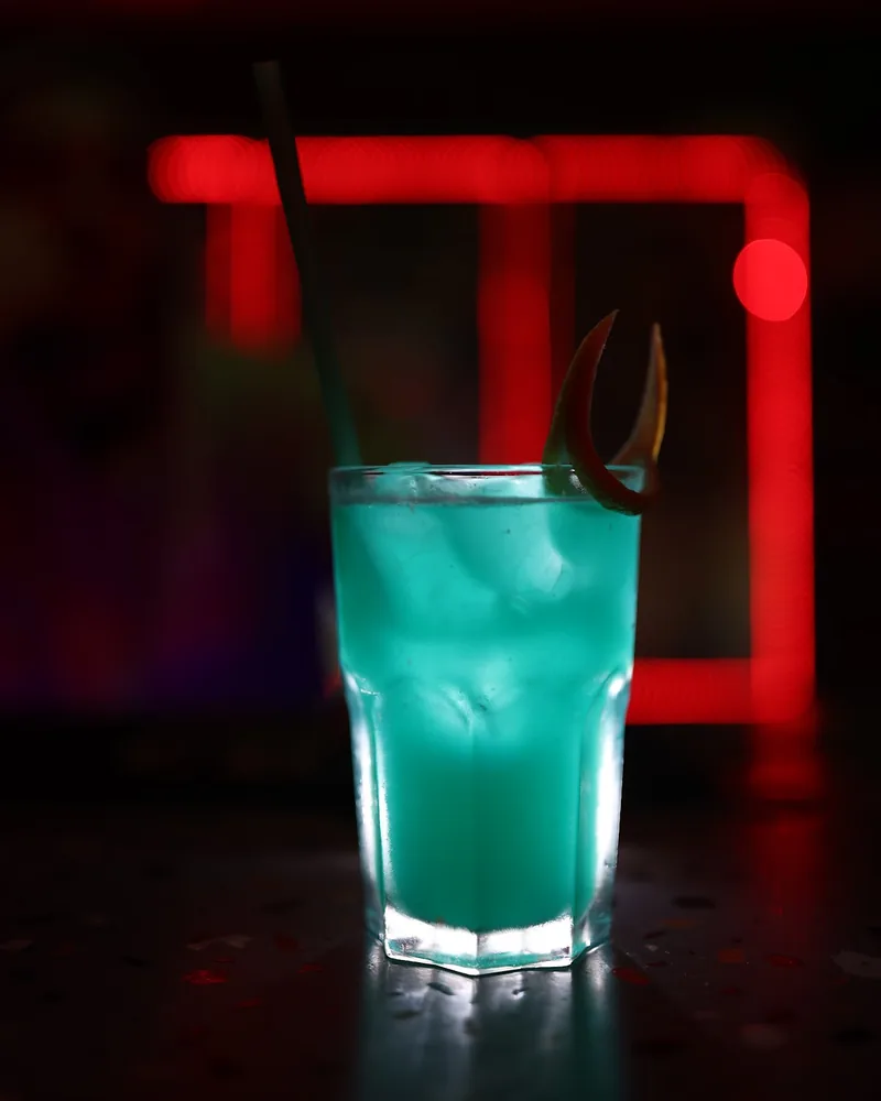 Cyan blue cocktail with peel garnish and straw in a tall glass at Ambavi