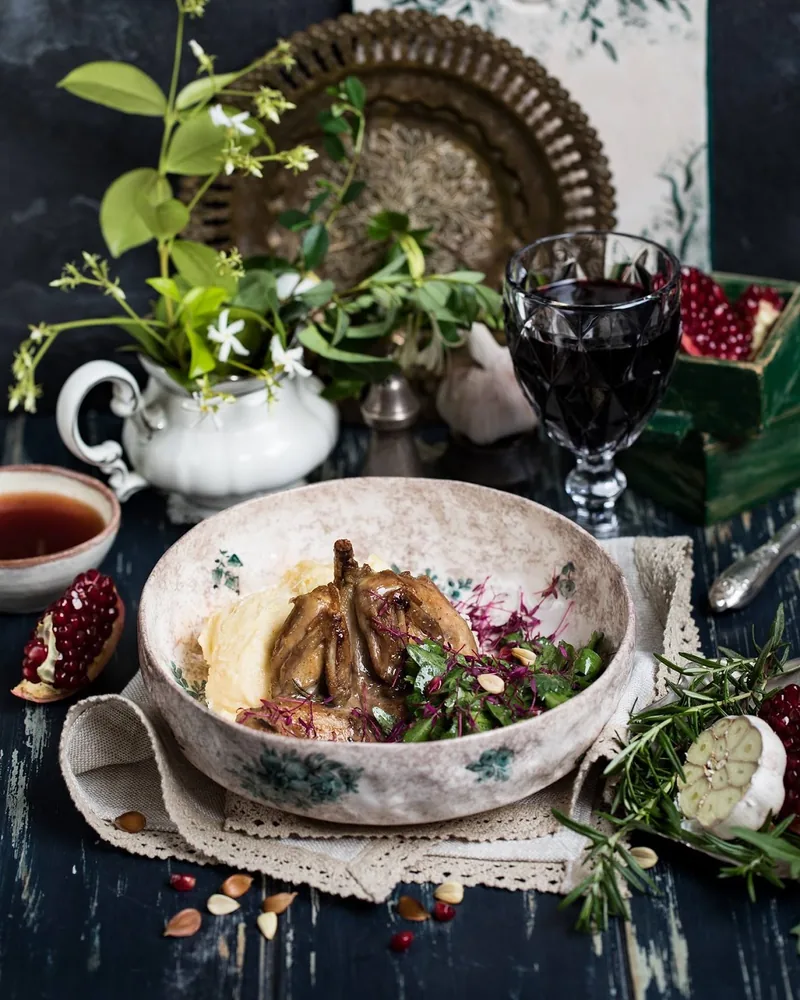 A roast quail on mashed potato with a herb salad in a patterned bowl, next to a glass of red wine and pomegranates, at Barbarestan