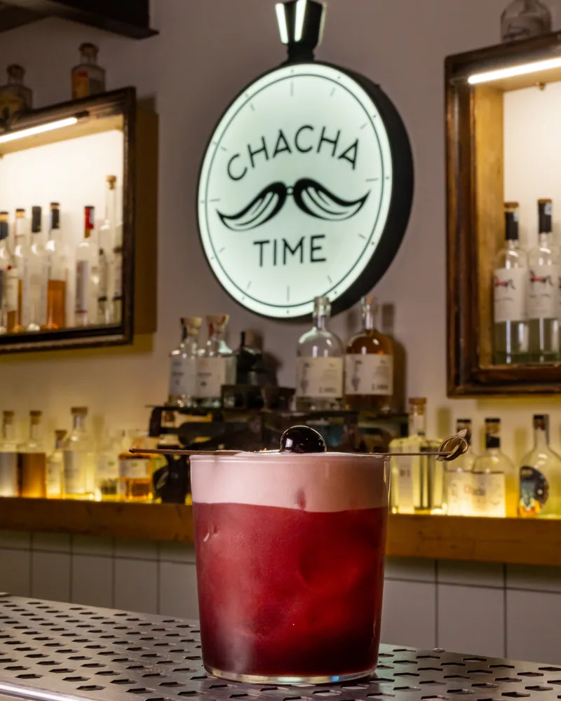 A red sour with a white foam top and a cherry on a pick, on a steel bar counter in front of the lit Chacha Time sign and shelves of bottles