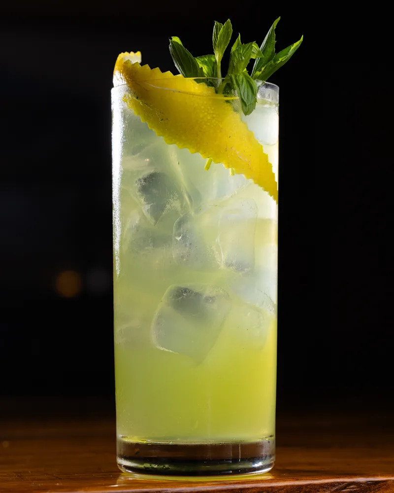 A pale green cocktail over ice in a highball glass with a lemon peel and mint at Chacha Time