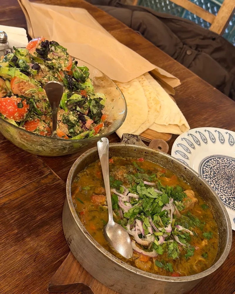 A metal pan of chashushuli stew topped with coriander and red onion, with a tomato salad and flatbread behind it