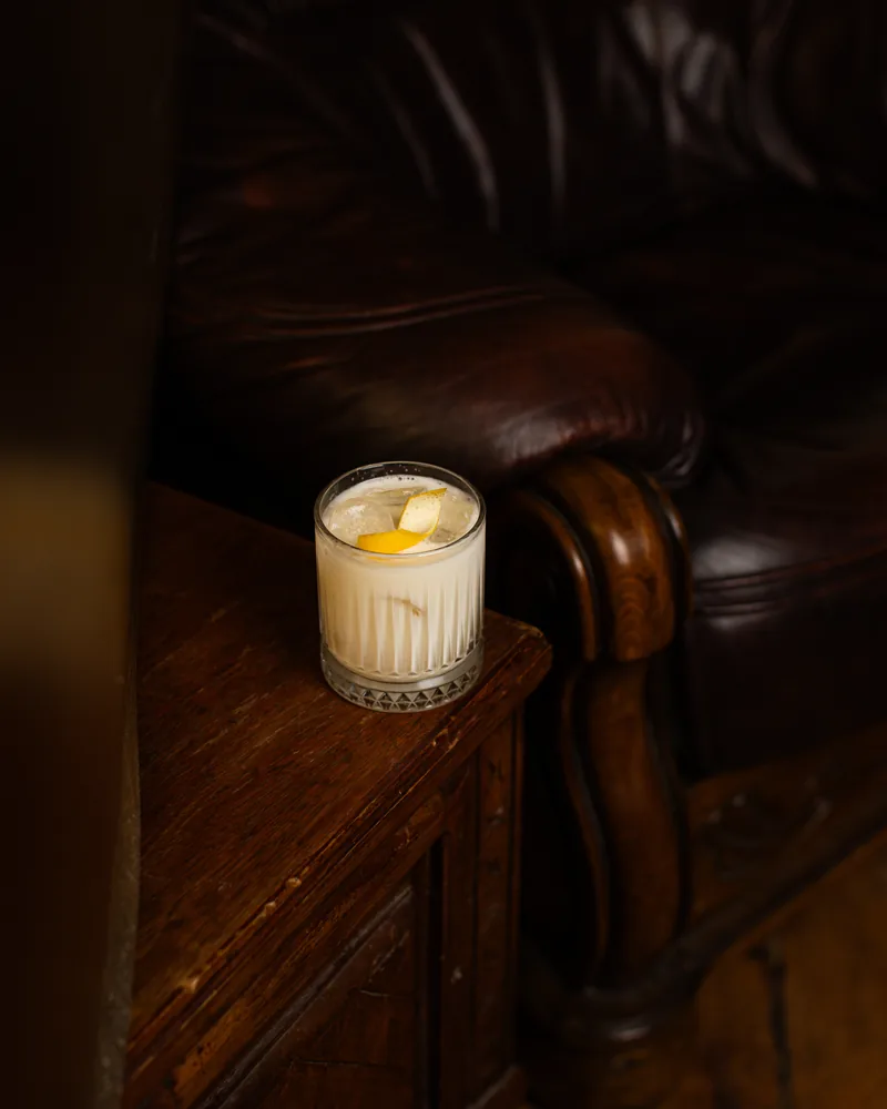 Pale creamy cocktail on ice with a lemon twist in a cut-crystal rocks glass, beside a leather armchair at Crossroads Bar