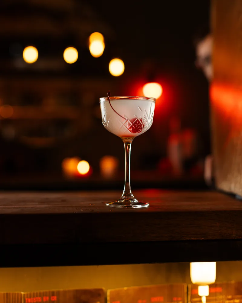 Frosted white cocktail with a stemmed cherry in a cut-crystal coupe on a wooden bar at Crossroads Bar