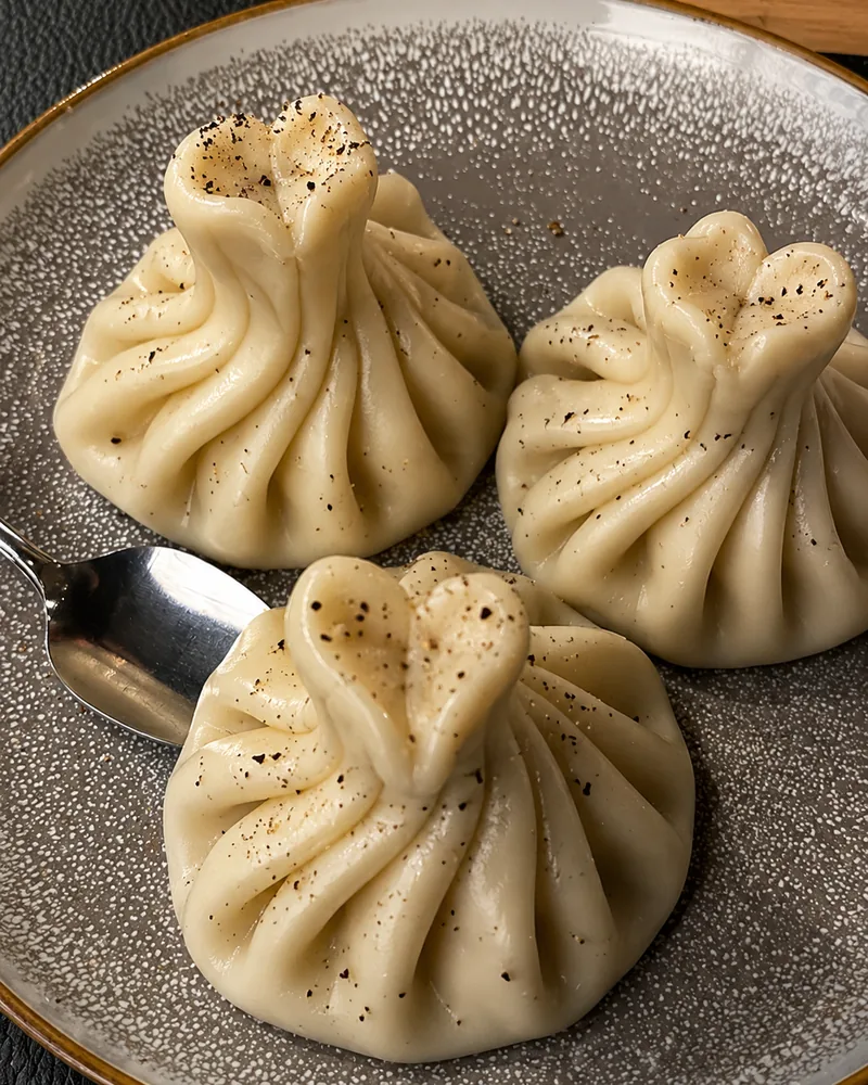 3 pleated khinkali dusted with black pepper on a grey speckled plate with a spoon, at Dariko Georgian Restaurant