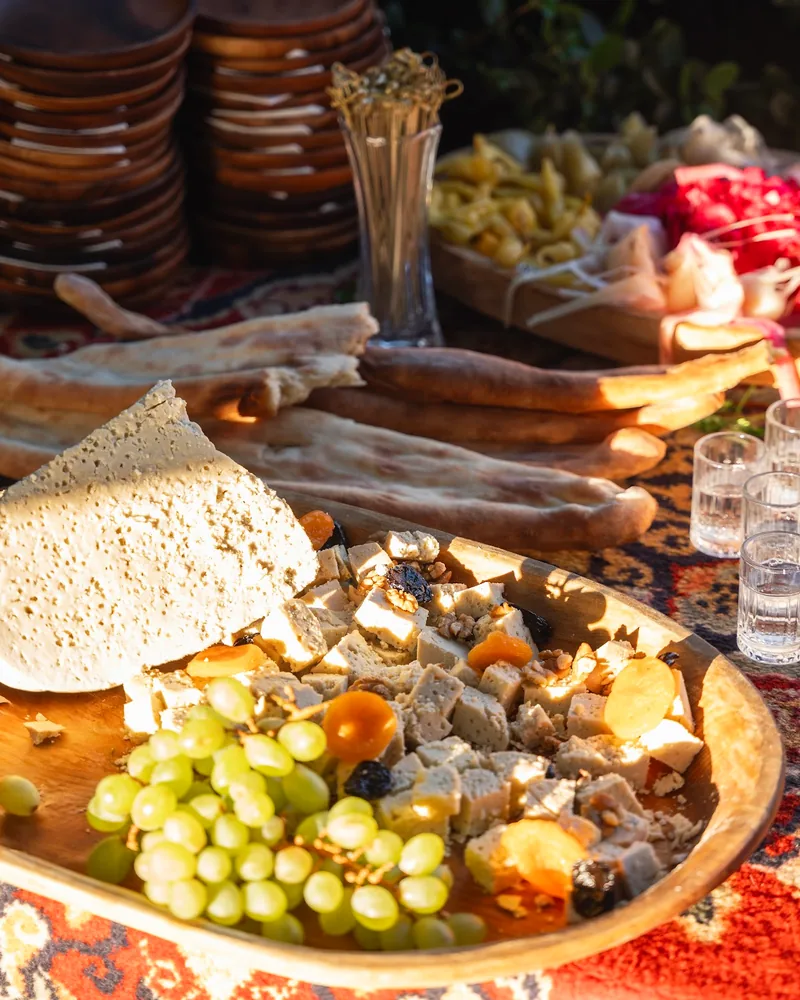 A wooden tray of cubed white cheese and a wedge of cheese full of small holes, with green grapes and dried apricots, long shotis puri loaves and small glasses behind