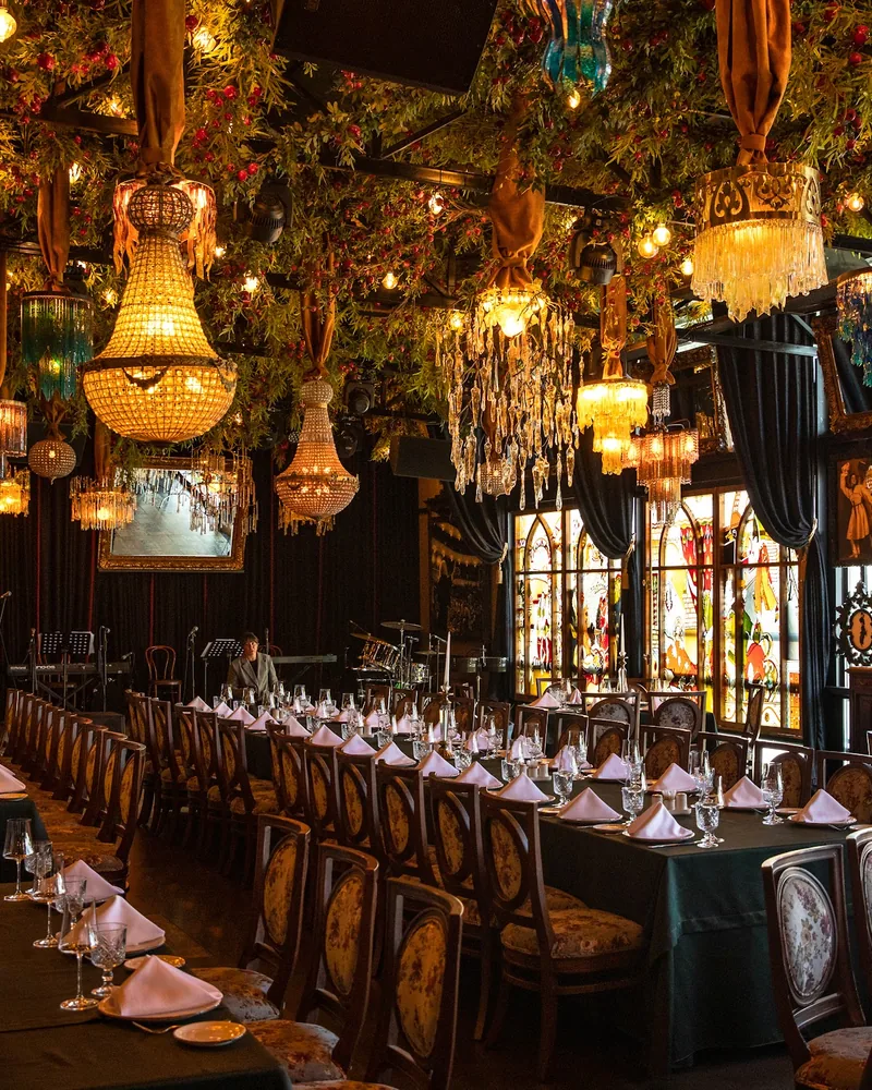 Long banquet tables with folded napkins under beaded chandeliers and hanging greenery, facing a stage with a drum kit, keyboards and microphone stands, at Ethnographer Restaurant