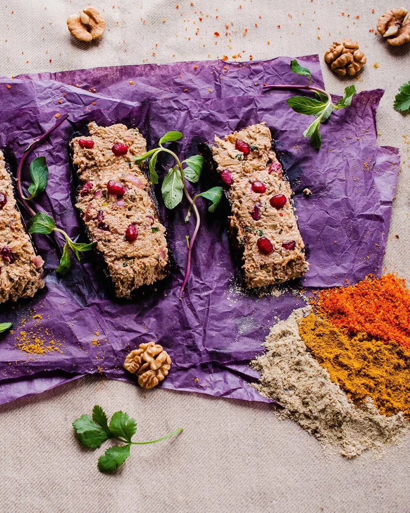 Fried aubergine slices spread with walnut paste and pomegranate seeds on purple paper, with walnuts and piles of ground spices, at Georgian Flavors
