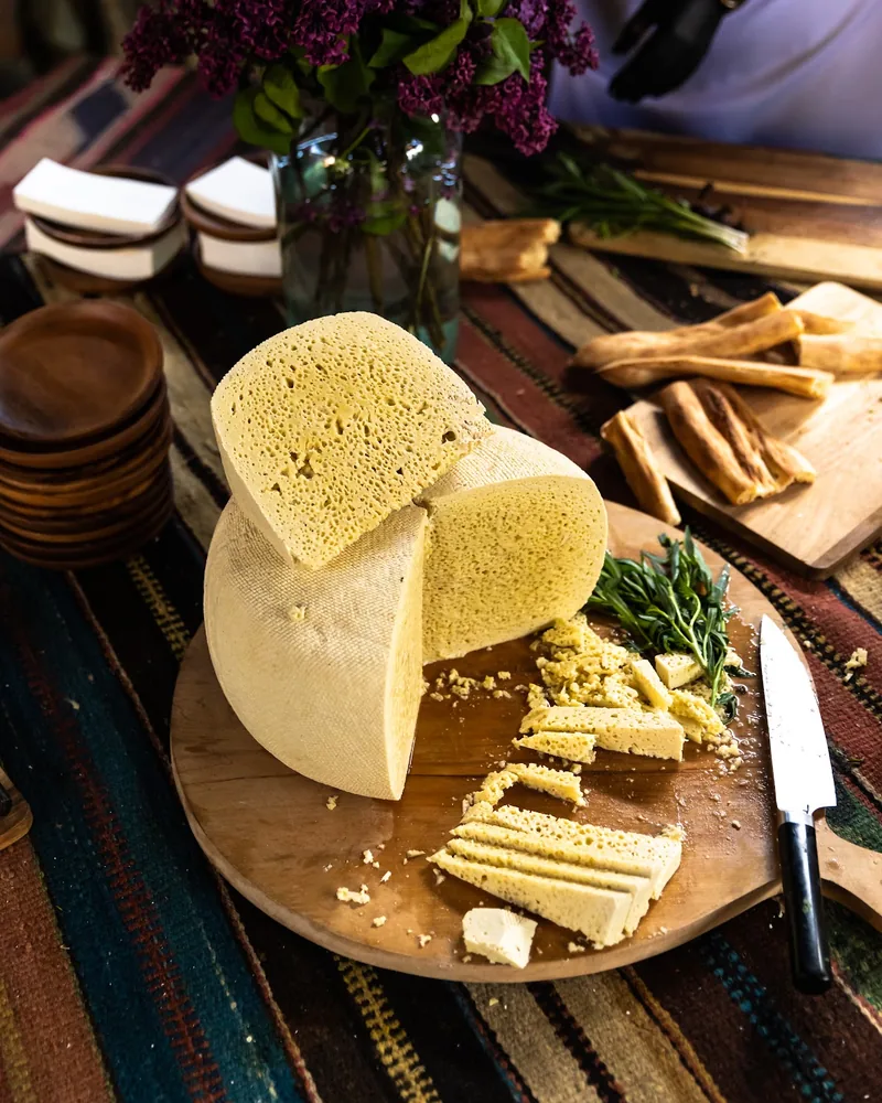 A wheel of pale yellow cheese with small holes, cut open on a round wooden board with slices, tarragon and bread sticks