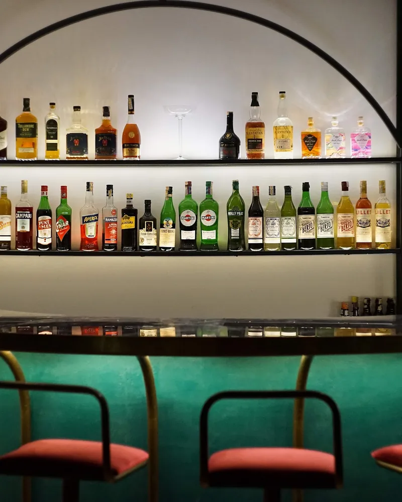 Backlit liquor shelves above curved green bar counter with red stools at Graali Bar