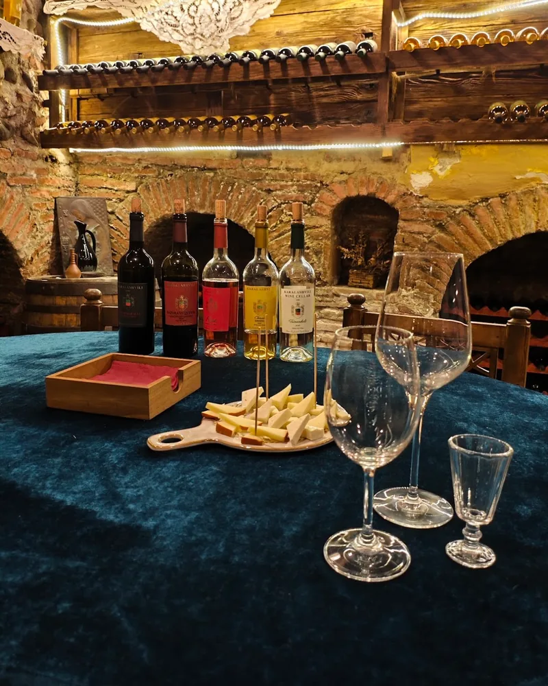A board of pale cheese cubes on wooden picks beside 5 bottles of wine, 2 wine glasses and a small glass on a velvet table in a brick-vaulted cellar
