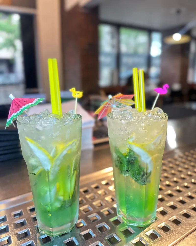 Two green cocktails with umbrellas and straws on a bar drip tray at Khushi Indian Restaurant