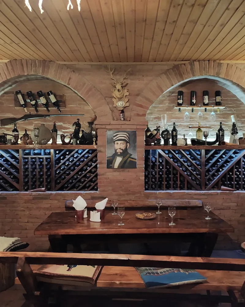 A long wooden table and benches under brick arches lined with wine racks, bottles and a painted portrait at Laliashvilebi's Wine Cellar