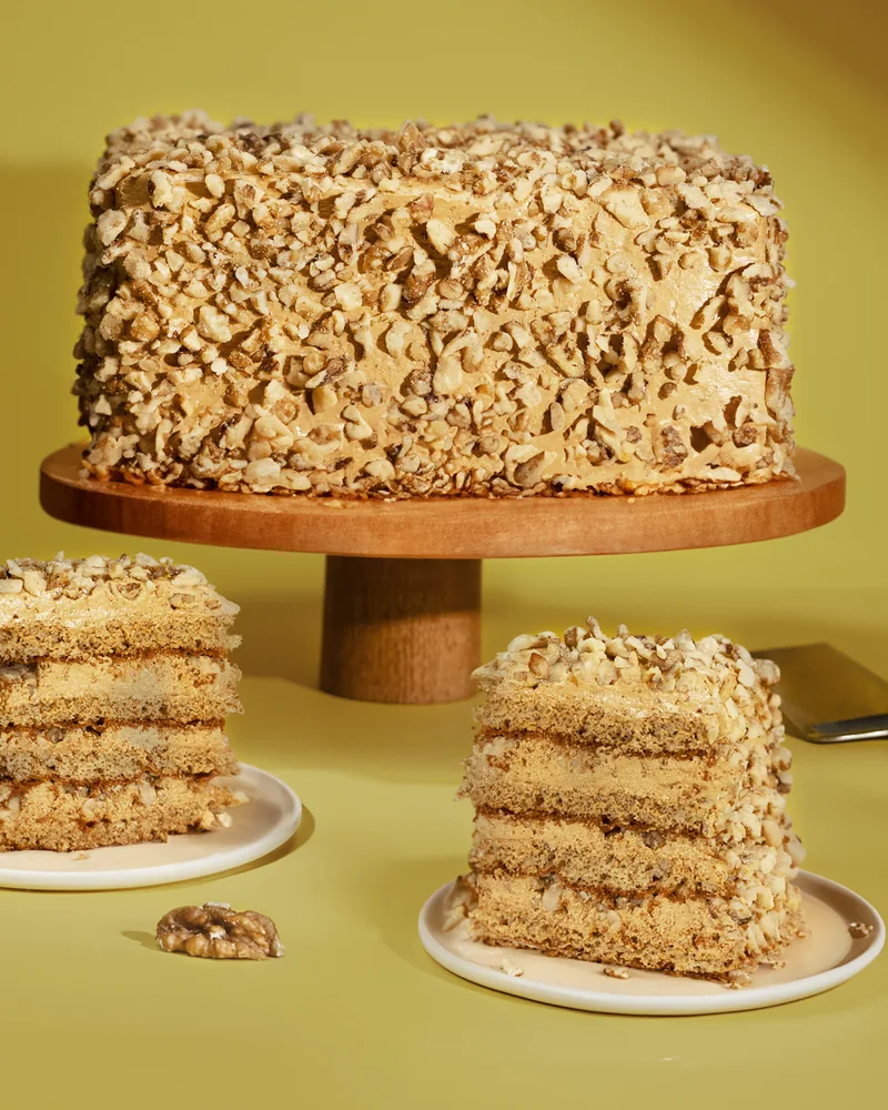 A walnut-crusted layer cake on a wooden cake stand with 2 cut slices on small plates