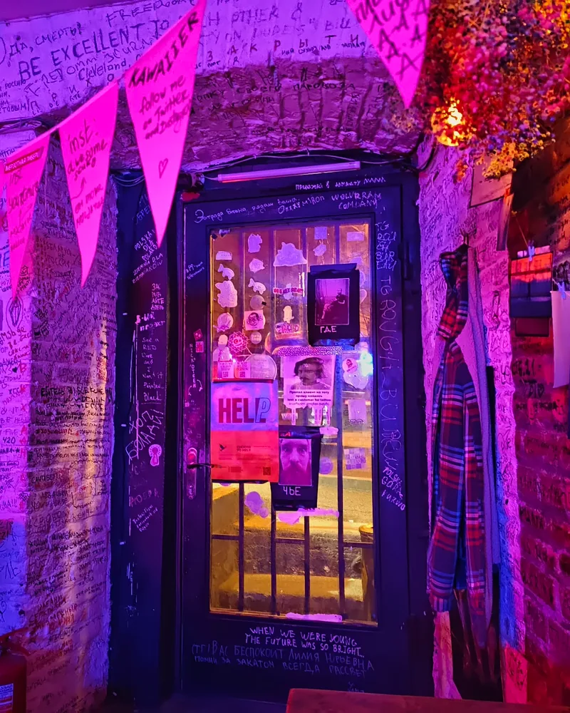 Graffiti-covered bar interior and entrance door under pink neon at Ploho Bar