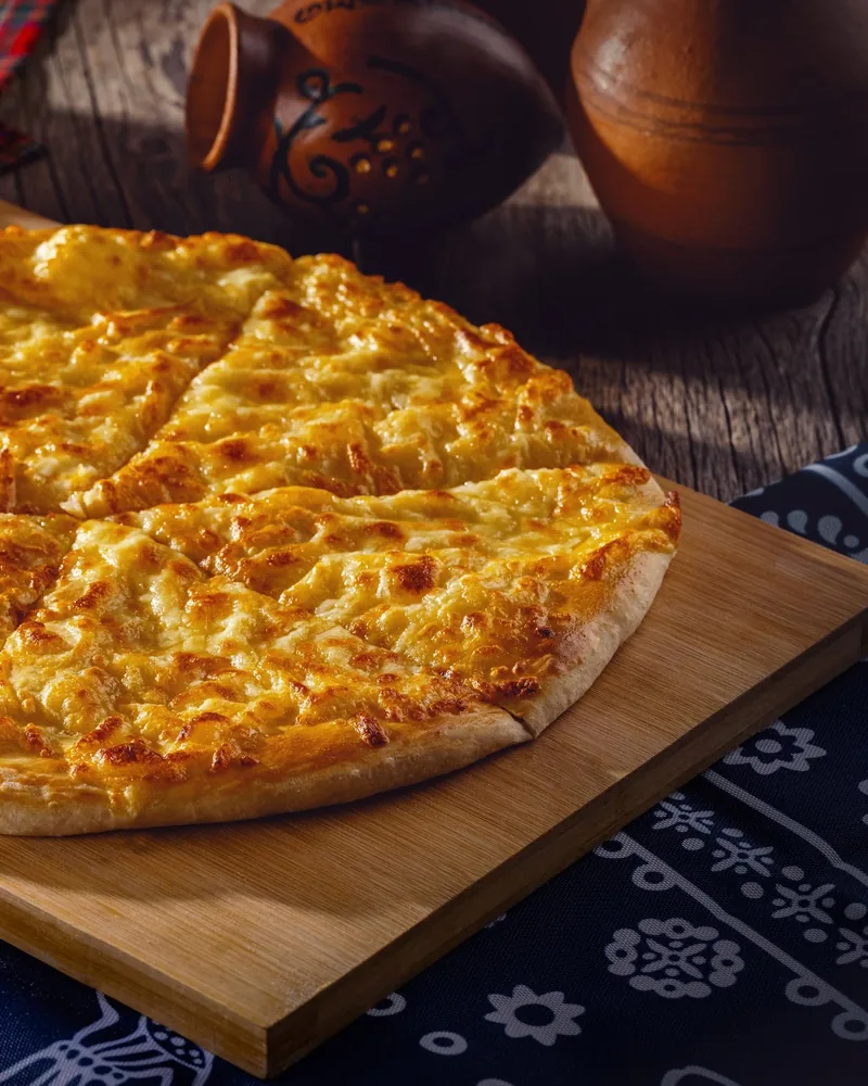 A Megrelian khachapuri with browned, blistered cheese baked across the top, cut into slices on a wooden board, with clay jugs behind