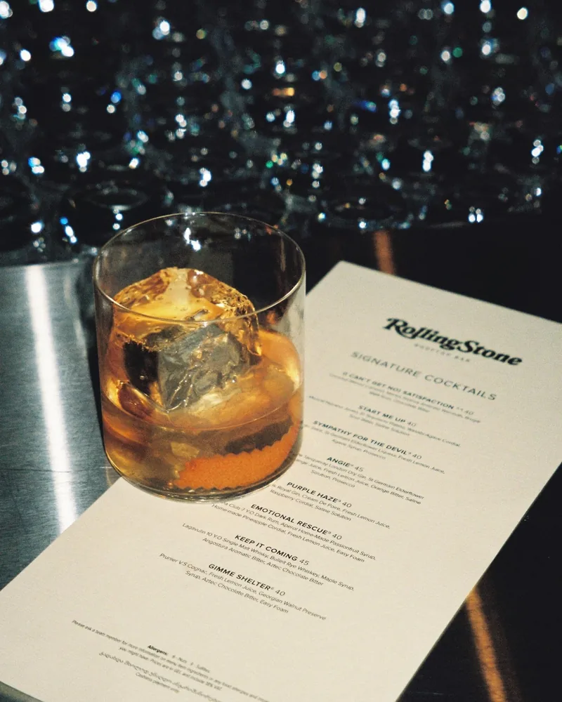 Cocktail with a large ice cube and orange peel beside the cocktail menu at Rolling Stone Rooftop Bar
