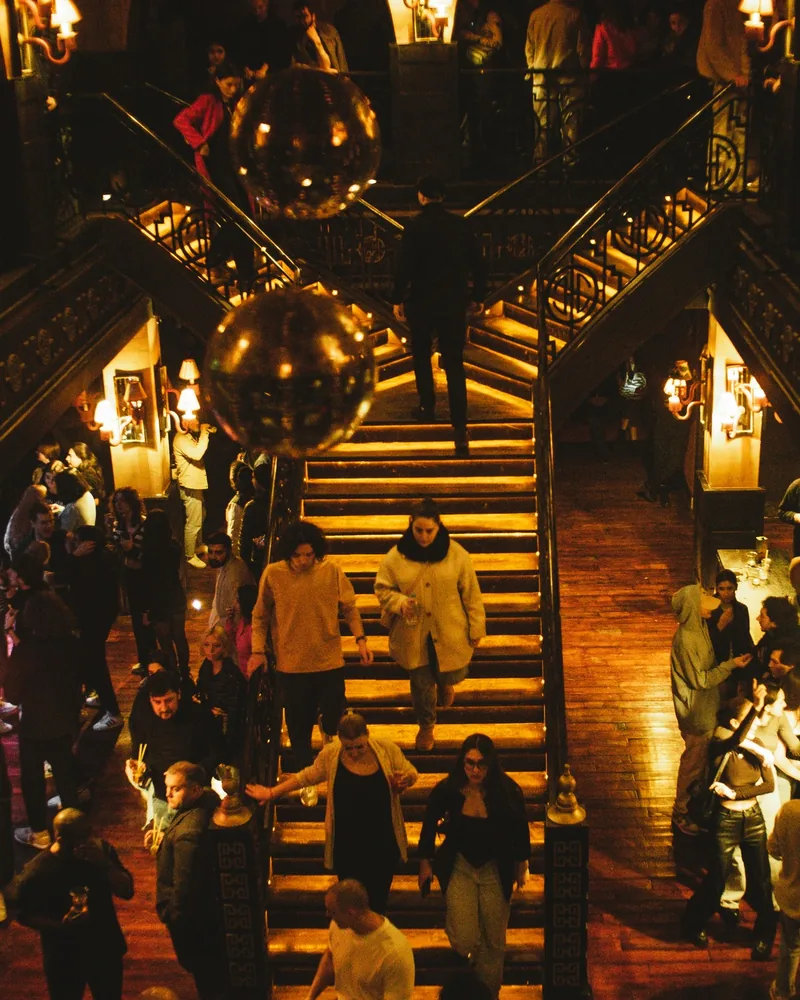 A 2-level bar room with a wide central staircase, ornate railings and 2 disco balls, with people on the stairs