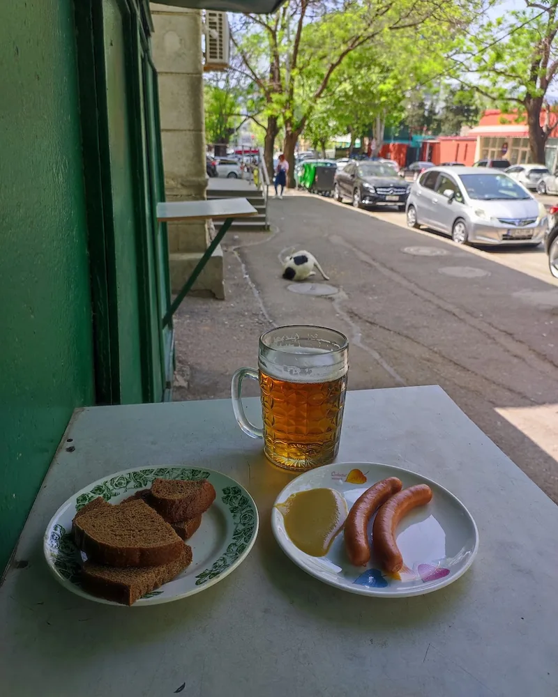A glass mug of draught beer, 2 sausages with yellow mustard and a plate of brown bread on a table by an open door, with a Tbilisi street behind