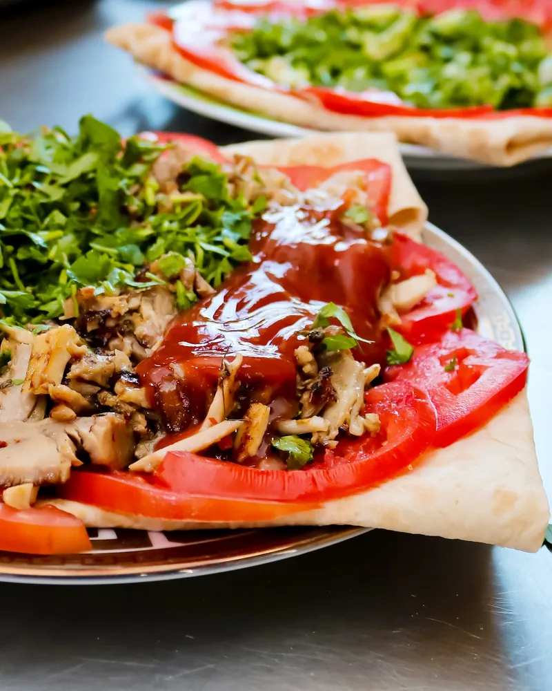 Shawarma laid open on lavash with sliced tomato, chopped herbs and a red sauce on a gold-rimmed plate