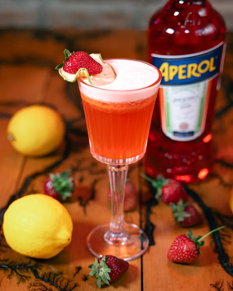 Cocktail in a stemmed glass with strawberry garnish and aperol bottle at So Lucky Cocktail Bar