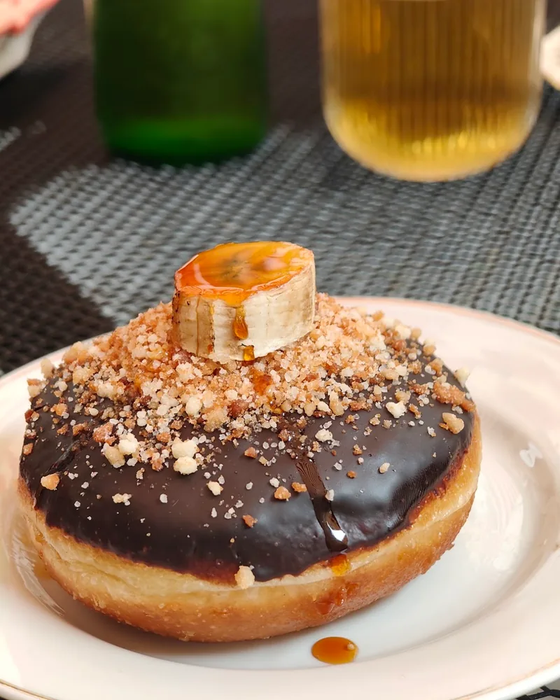 A chocolate-glazed doughnut topped with crumble and a slice of caramelised banana on a white plate
