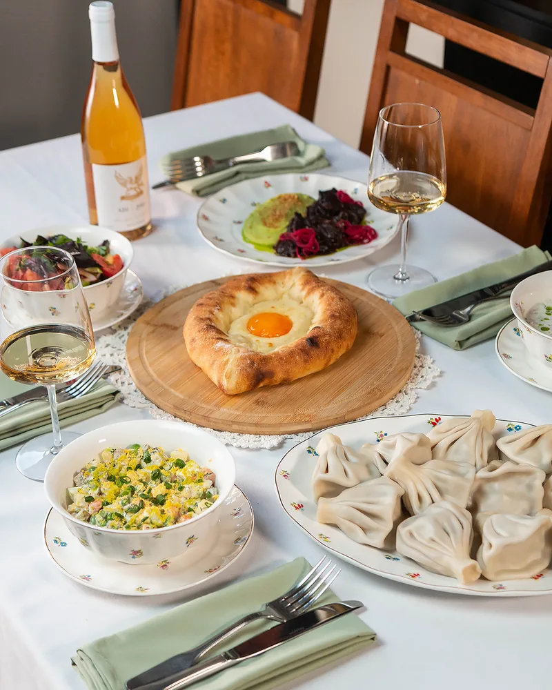 An Adjarian khachapuri with an egg yolk on a wooden board, a platter of khinkali, salads, a bottle of amber wine and 2 glasses on a white tablecloth at Stumari