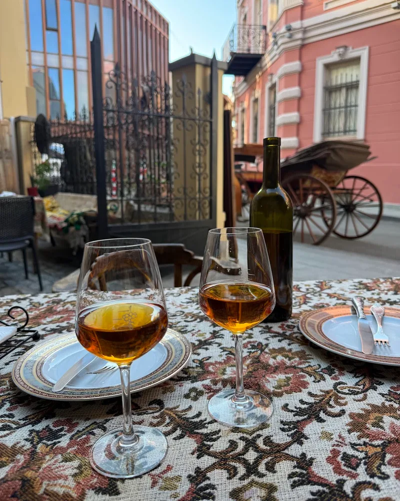 2 glasses of amber wine, one deeper than the other, and a bottle on a patterned tablecloth at an outdoor table on an old Tbilisi street