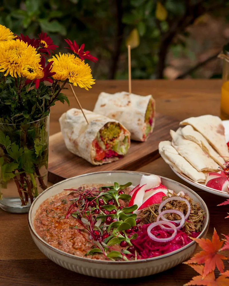 A bowl of lobio topped with microgreens and pomegranate seeds, with pickled red cabbage, radishes and onion rings, and a halved wrap behind