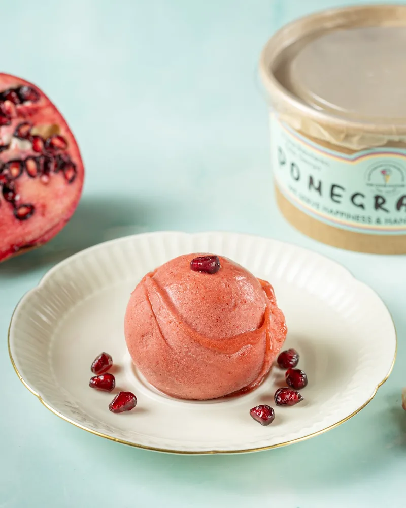 A scoop of pink pomegranate ice cream with pomegranate seeds on a white plate, a cut pomegranate beside it
