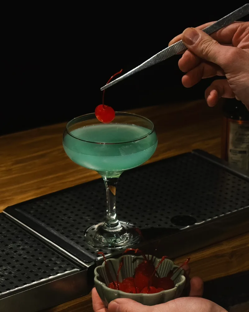 A pale blue cocktail in a coupe on a bar mat, with a cherry being lowered in by tweezers, at The Gin Corner