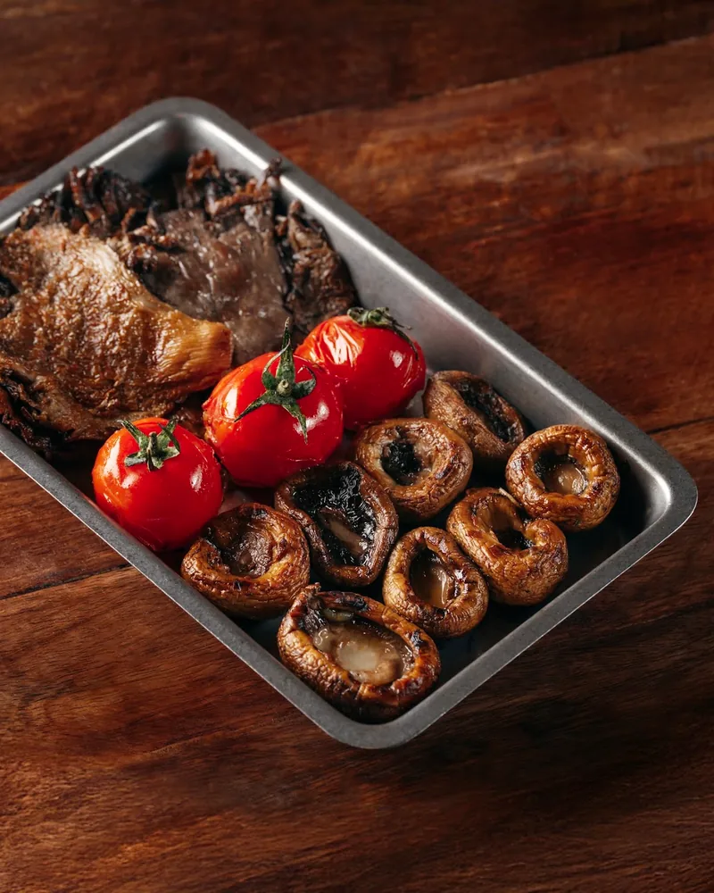 Grilled meat with blistered cherry tomatoes and roasted mushrooms in a metal tray on a wooden table