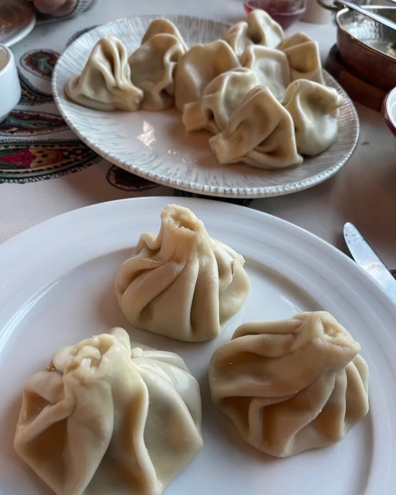 3 boiled khinkali on a white plate, with a platter of more khinkali behind them