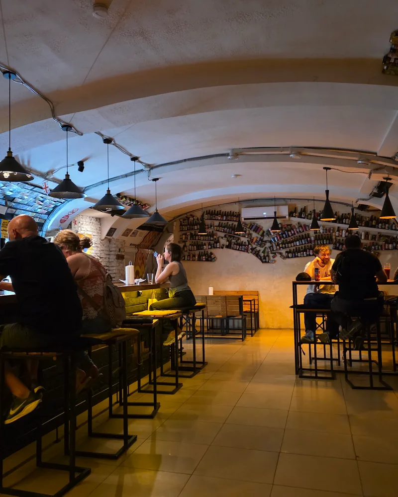 A low vaulted room with high tables, black pendant lamps and a back wall lined with beer bottles