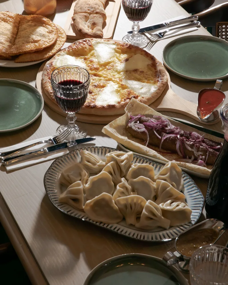 A platter of khinkali, a cheese khachapuri, a kebab on flatbread with red onion and glasses of red wine on a wooden table at Vakhtanguri