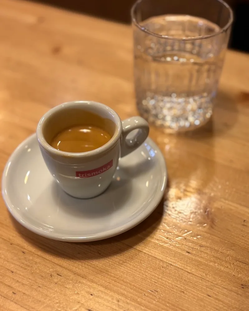 An espresso in a white cup on a saucer, with a glass of water behind it, on a wooden table at Walkers Espresso Bar