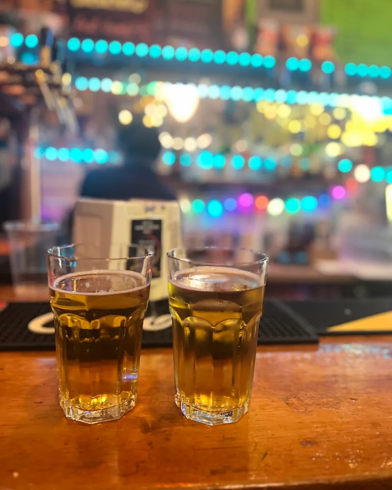 2 glasses of pale lager on a wooden bar counter under strings of coloured lights