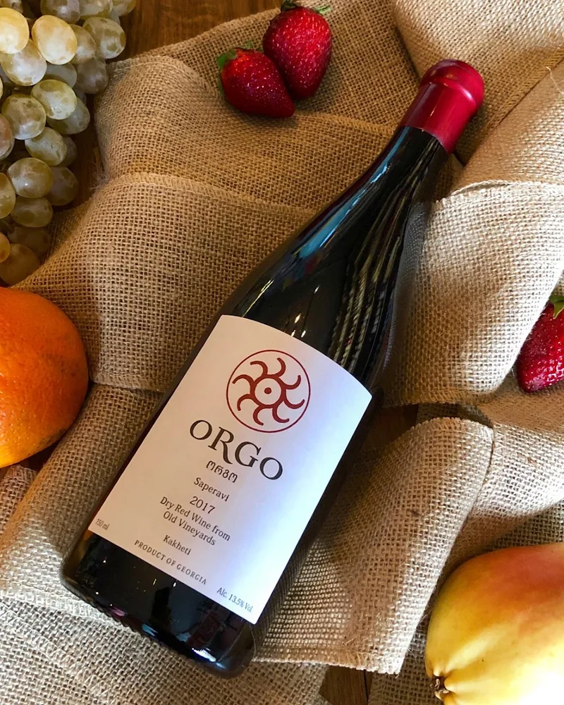 A bottle of Orgo Saperavi 2017, a dry red from old vineyards in Kakheti, lying on burlap with white grapes, strawberries, an orange and a pear